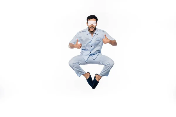 Uomo stupito in pigiama blu e maschera addormentata levitante mentre mostra i pollici in su sul bianco — Foto stock