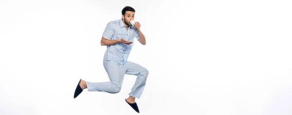 Hombre barbudo en pijama sosteniendo la taza y bebiendo café mientras levita en blanco, pancarta - foto de stock