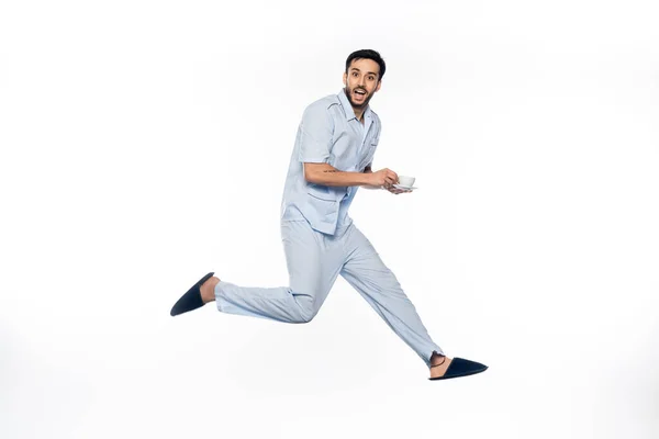 Hombre feliz en pijama sosteniendo taza y platillo mientras levita sobre blanco - foto de stock