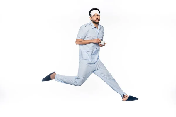 Bärtiger Mann im Pyjama hält Tasse und Untertasse in der Hand, während er auf Weiß schwebt — Stockfoto