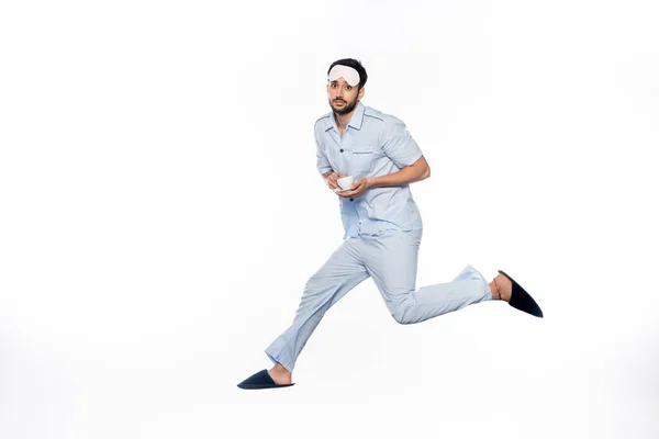 Homme barbu en pyjama et masque de couchage tenant tasse avec soucoupe tout en lévitant sur blanc — Photo de stock