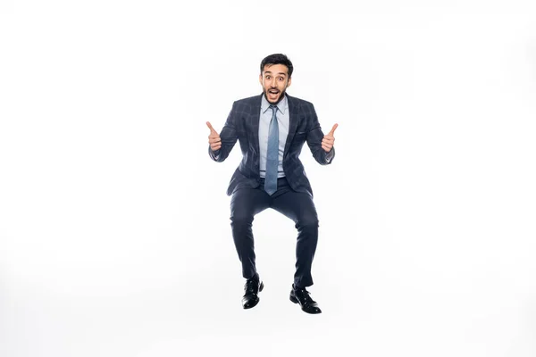 Excitado hombre de negocios en traje saltando mientras muestra los pulgares hacia arriba en blanco - foto de stock