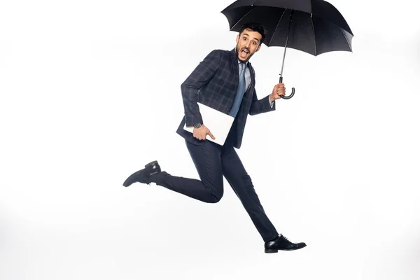 Возбужденный бизнесмен в костюме прыгать с ноутбука и зонтиком на белом — стоковое фото