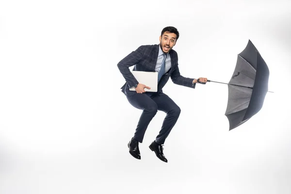 Stupito uomo d'affari in tuta che salta con computer portatile e ombrello su bianco — Foto stock