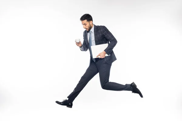Шокований бізнесмен в костюмі стрибає, тримаючи ноутбук і чашку на білому — стокове фото