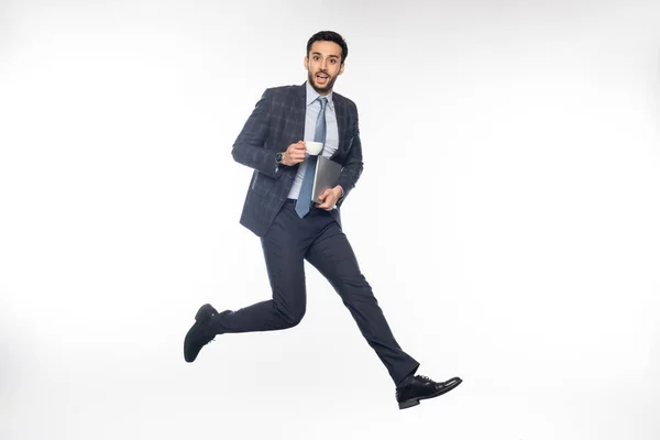 Empresário animado em terno pulando enquanto segurando laptop e xícara no branco — Fotografia de Stock