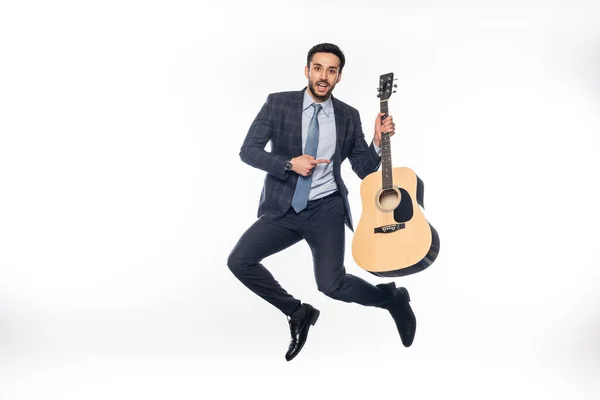 Happy businessman in suit jumping and pointing at acoustic guitar on white — Stock Photo