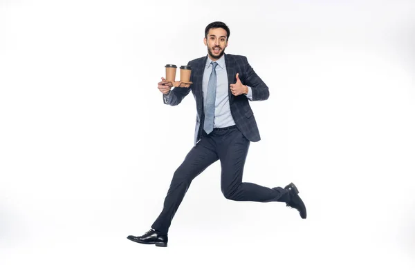 Happy businessman in suit jumping while holding carton cup holder with paper cups and showing thumb up on white — Stock Photo