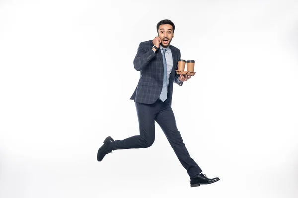 Hombre de negocios barbudo en traje saltando mientras sostiene el soporte de la taza de cartón con copas de papel y hablando en el teléfono inteligente en blanco - foto de stock