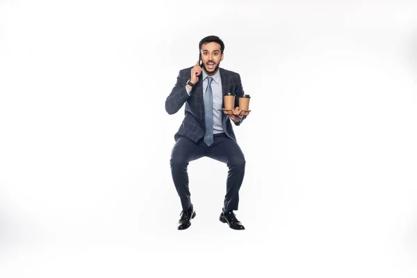 Happy businessman in suit jumping while holding cup holder with paper cups and talking on smartphone on white — Stock Photo