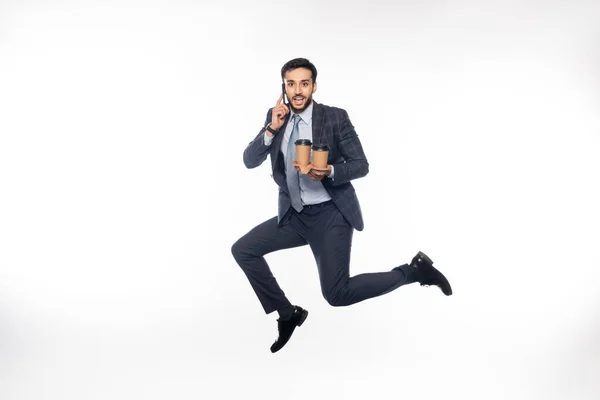 Cheerful businessman in suit jumping while holding cup holder with paper cups and talking on smartphone on white — Stock Photo