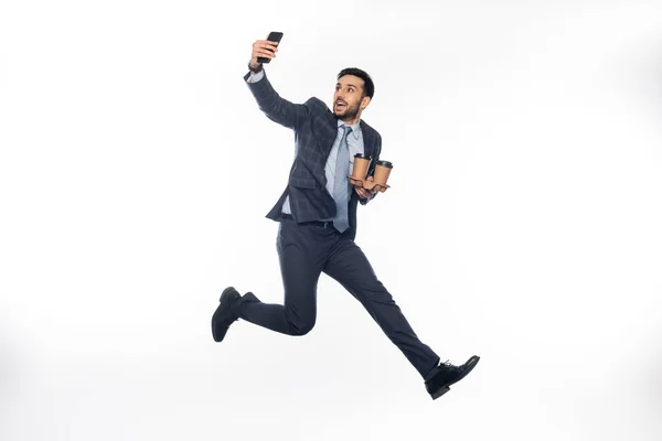 Cheerful businessman in suit jumping while holding cup holder with paper cups and taking selfie on white — Stock Photo