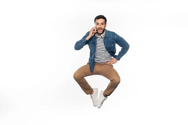 Homem feliz levitando enquanto fala no smartphone e olhando para a câmera no branco — Fotografia de Stock