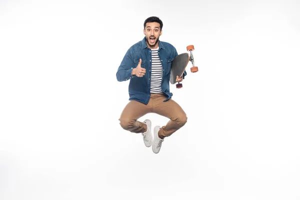 Homem feliz levitar enquanto segurando longboard e mostrando polegar para cima no branco — Fotografia de Stock