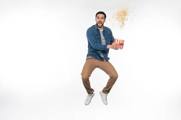 Hombre excitado levitando mientras sostiene cubo cerca de palomitas de maíz voladoras en blanco - foto de stock