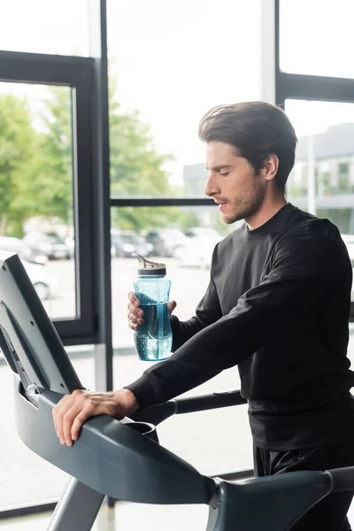 Vue latérale du sportif tenant une bouteille de sport sur tapis roulant dans la salle de gym — Photo de stock