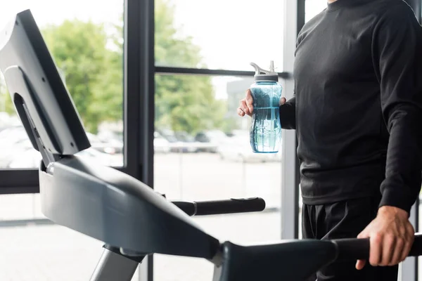 Vista ritagliata dello sportivo che tiene la bottiglia sportiva mentre si allena sul tapis roulant in palestra — Foto stock