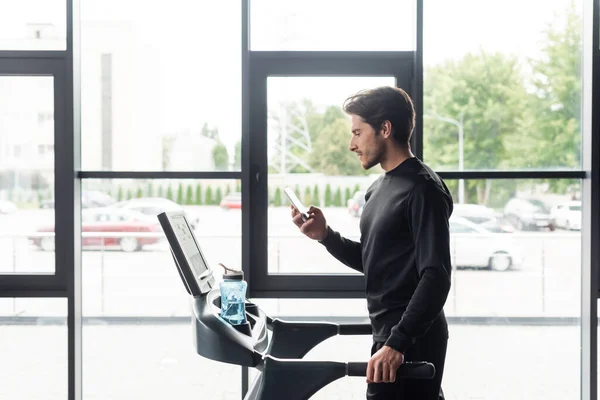 Vue latérale du sportif utilisant un smartphone pendant l'entraînement sur tapis roulant dans la salle de gym — Photo de stock