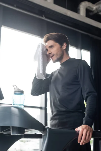 Спортсмен держит полотенце во время тренировки на беговой дорожке возле спортивной бутылки в тренажерном зале — стоковое фото