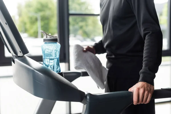 Обрезанный вид спортсмена, держащего размытое полотенце во время тренировки на беговой дорожке со спортивной бутылкой в тренажерном зале — стоковое фото