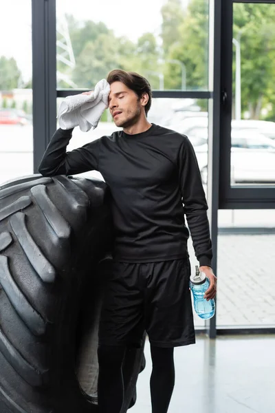 Brunette sportsman wiping face with towel and holding sports bottle near tire in gym — Foto stock