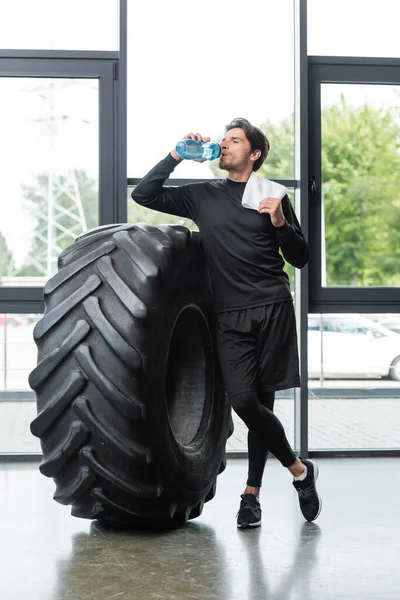 Sportsman drinking water and holding towel near tire in sports center — Stock Photo