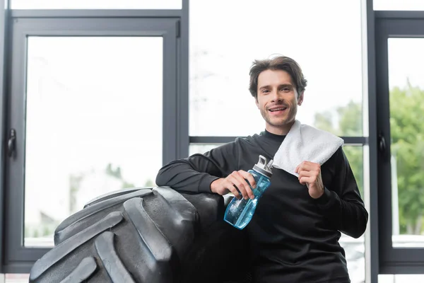 Deportista sonriente sosteniendo botella de deporte y toalla cerca de neumático en el gimnasio - foto de stock