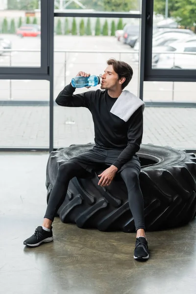 Brunette sportsman with towel drinking water while sitting on tire in sports center — Stockfoto