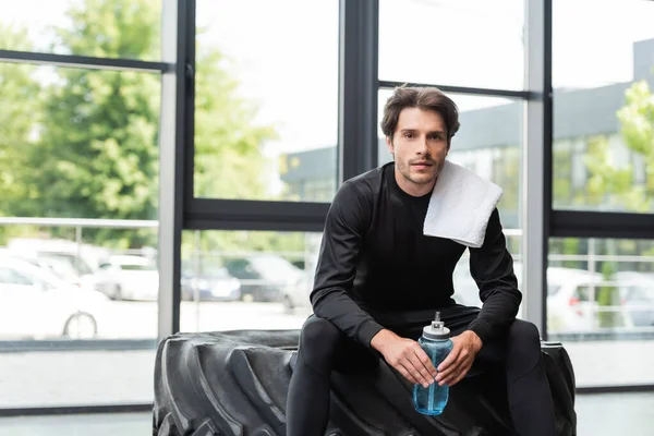 Sportsman avec serviette et bouteille de sport assis sur le pneu dans la salle de gym — Photo de stock