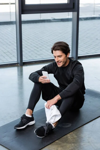 Sportif souriant utilisant un smartphone et tenant une serviette sur un tapis de fitness dans une salle de gym — Photo de stock