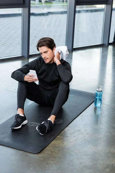 Sportsman using smartphone and wiping neck with towel on fitness mat in gym — Stock Photo