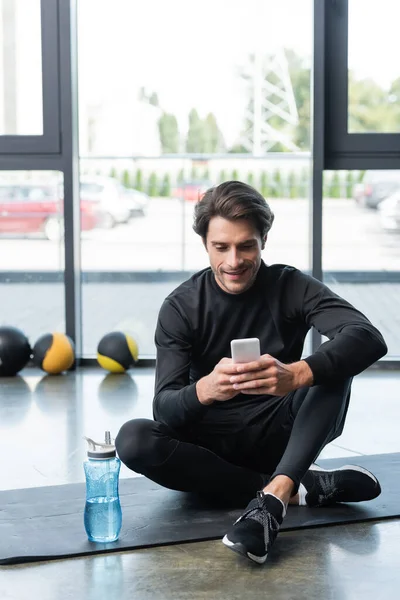 Esportista alegre usando celular perto de garrafa de esportes no tapete de fitness no ginásio — Fotografia de Stock