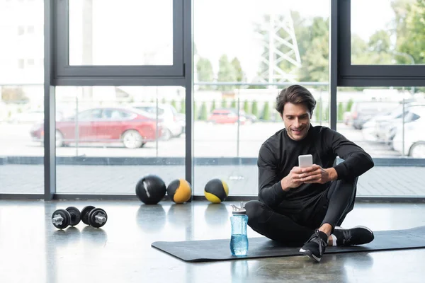 Positive sportsman using smartphone near sports bottle on fitness mat and dumbbells in gym — Stock Photo