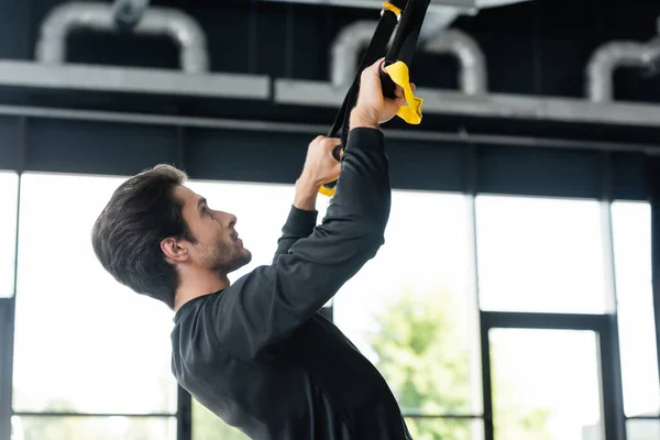 Side view of brunette sportsman pulling suspension straps in sports center — Stockfoto