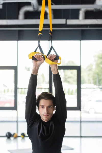 Brunette sportsman training with suspension straps and looking at camera in gym — Foto stock