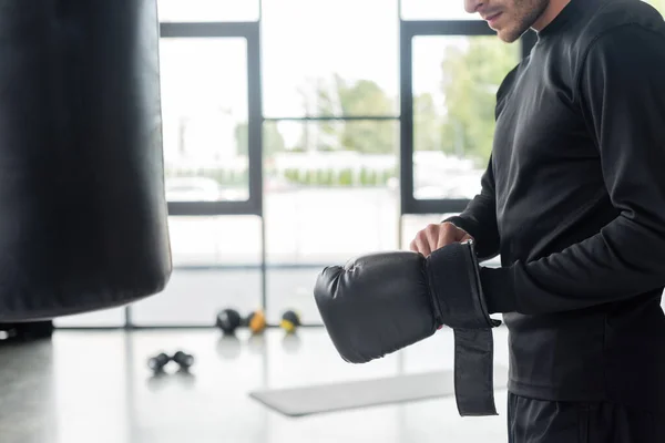 Cropped view of sportsman wearing boxing glove near punching bag in gym — Stock Photo