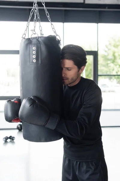 Спортсмен в боксёрских перчатках обнимает боксерскую грушу в спортзале — стоковое фото