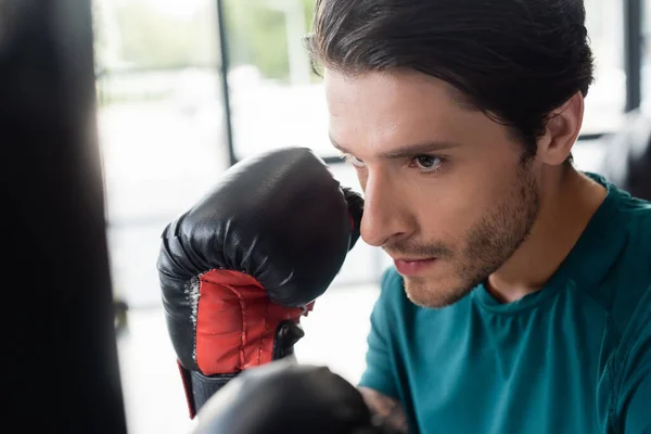 Retrato de desportista em luvas de boxe olhando para saco de perfuração borrada no ginásio — Fotografia de Stock
