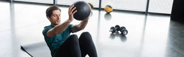 Брюнетка спортсмен тренирующийся с мячом возле гантелей в тренажерном зале, баннер — стоковое фото