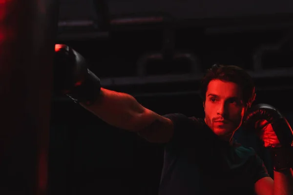 Sportsman in boxing gloves training with punching bag on black background with red light — Stock Photo