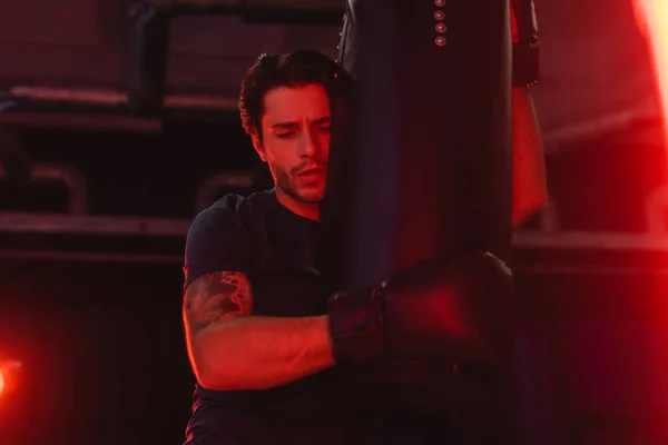 Sportsman hugging punching bag in gym with red light — Stock Photo