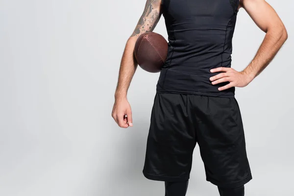 Обрезанный вид спортсмена с мячом для регби, держащего руку на бедре на сером фоне — стоковое фото