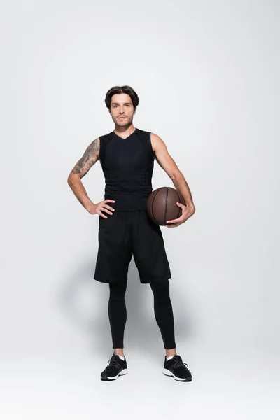 Full length of tattooed player with basketball ball looking at camera on grey background — Stockfoto