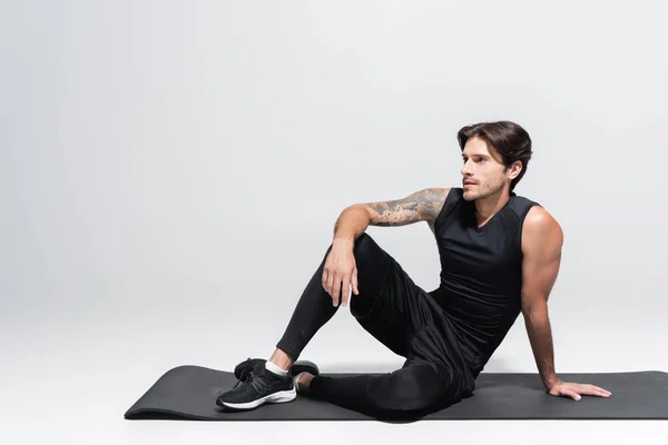 Sportsman sitting on black fitness mat on grey background — Stock Photo