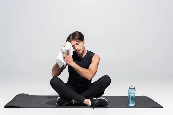 Sportsman essuyant visage avec serviette près de bouteille de sport sur tapis de fitness sur fond gris — Photo de stock