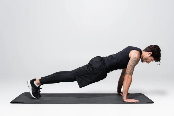 Side view of sportsman in back sportswear doing plank on fitness mat on grey background — Stockfoto