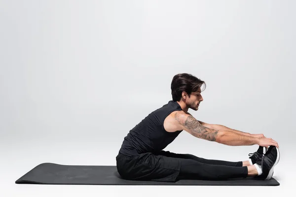 Vue latérale du sportif brunette s'échauffant sur tapis de fitness sur fond gris — Photo de stock