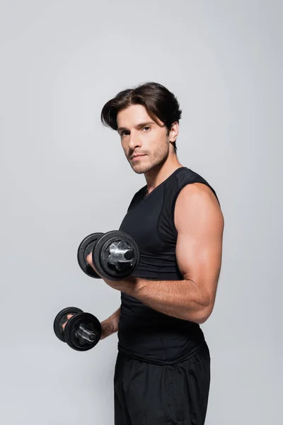 Hombre atlético en ropa deportiva sosteniendo mancuernas y mirando a la cámara aislada en gris — Stock Photo