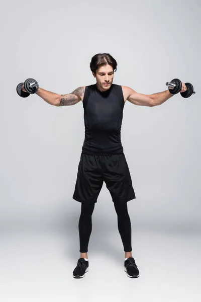 Full length of tattooed sportsman training with dumbbells on grey background — Stock Photo