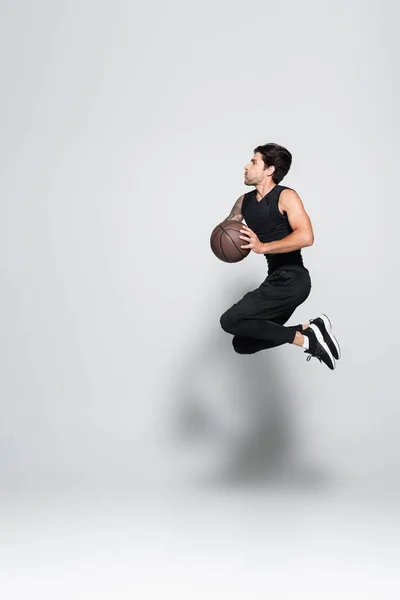 Vista lateral do desportista tatuado com bola de basquete saltando sobre fundo cinza — Fotografia de Stock